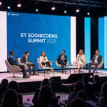 A dynamic eye-level shot of a diverse panel of speakers on a well-lit stage at a professional business summit. The large LED screen behind them displays the words "ET Soonicorns Summit 2025." Six speakers are seated at a table, engaged in a discussion. The stage is modern and the foreground shows the backs of an attentive audience.