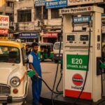A man is filling a white and yellow car with fuel at a gas station in India. The fuel pump has an "E20 ETHANOL BLEND" sign on it. There are other cars and auto-rickshaws in the background. The scene is set in a city.